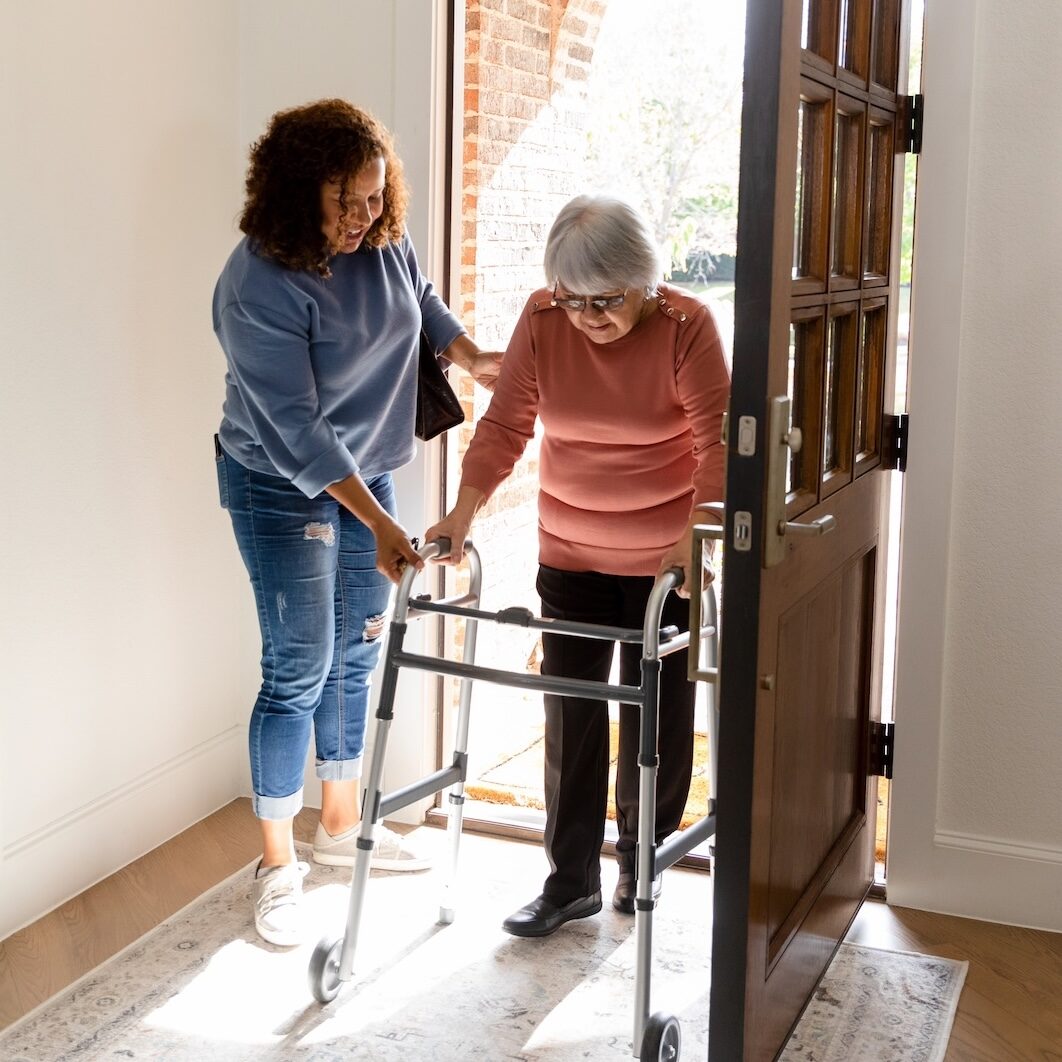 The mid adult woman helps her senior adult friend carefully enter her home.