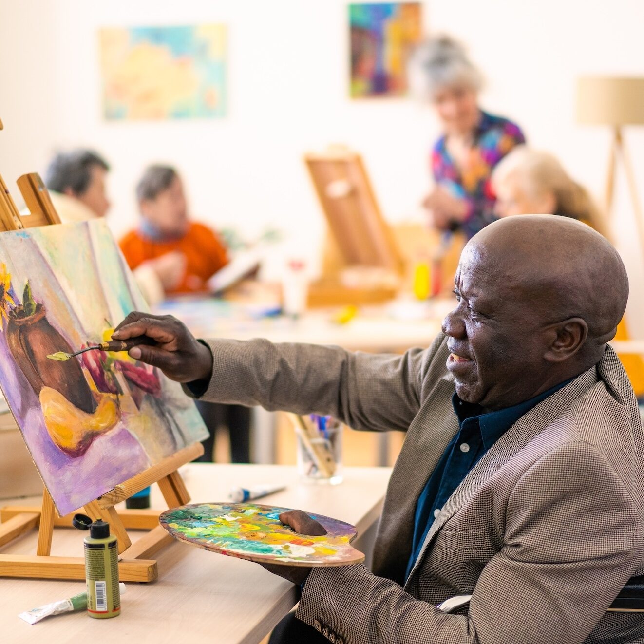 A retired Anglo-Caribbean man is painting a picture onto a canvas supported on an easel, holding a palette in one hand, behind more senior people are also painting